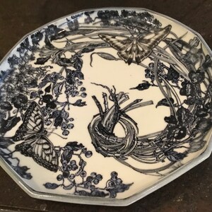 May include: A decorative ceramic plate with a cream-colored center and a blue and white butterfly and floral design. The plate has an irregular, octagonal shape with a blue border. The design includes butterflies, berries, and foliage.