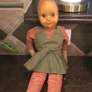 Może przedstawiać: Zabytkowa lalka z malowaną twarzą, ubrana w zieloną sukienkę i czerwono-białe legginsy w paski. Lalka ma materiałowy korpus i malowaną twarz z dużymi niebieskimi oczami. Ręce lalki pokryte są czerwono-białą tkaniną w paski.