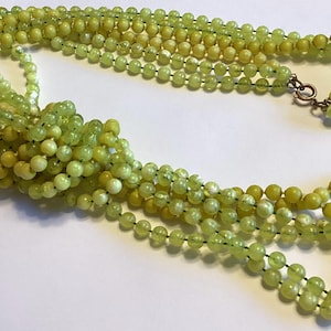 May include: A multi-strand necklace with small, round, light green beads. The beads are strung together with dark thread and secured with a copper-coloured clasp. The necklace is knotted at the base, creating a layered effect.