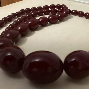 Op de afbeelding: Een ketting met gegradueerde kralen in een gladde, donkerbordeauxrode kleur, vari&euml;rend in ronde en ovale vormen. De kralen worden groter naar het midden van de streng toe, die aan het uiteinde is voorzien van een zilverkleurige metalen sluiting.
