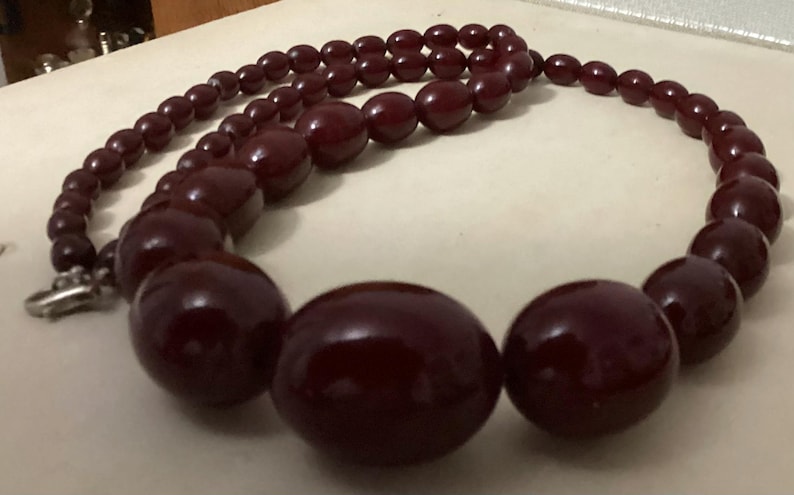 Op de afbeelding: Een ketting met gegradueerde kralen in een gladde, donkerbordeauxrode kleur, vari&euml;rend in ronde en ovale vormen. De kralen worden groter naar het midden van de streng toe, die aan het uiteinde is voorzien van een zilverkleurige metalen sluiting.