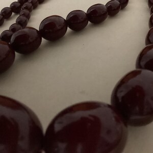 Op de afbeelding: Een ketting met een streng gladde, gepolijste, donkerbordeauxrode kralen. De kralen zijn in grootte gegradueerd, met grotere, afgeronde, ovaalvormige kralen in het midden en kleinere, ronde kralen langs de zijkanten van de streng.