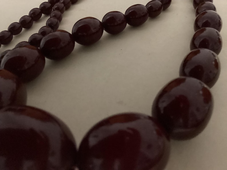Op de afbeelding: Een ketting met een streng gladde, gepolijste, donkerbordeauxrode kralen. De kralen zijn in grootte gegradueerd, met grotere, afgeronde, ovaalvormige kralen in het midden en kleinere, ronde kralen langs de zijkanten van de streng.