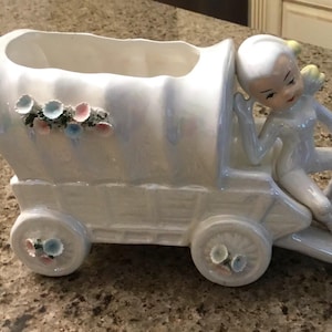 May include: A white ceramic wagon planter with a seated cherub figure. The wagon has four wheels and is decorated with floral accents in pink, blue, and white. The cherub has a serene expression.