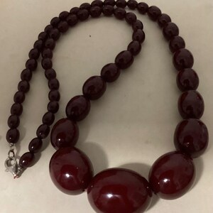 Op de afbeelding: Een ketting met gegradueerde kralen bestaande uit gepolijste, diep bordeauxrode ovale kralen. De kralen worden groter naar het midden toe, waar de grootste ovale kraal is geplaatst. De ketting wordt gesloten met een zilverkleurige metalen karabijnsluiting.