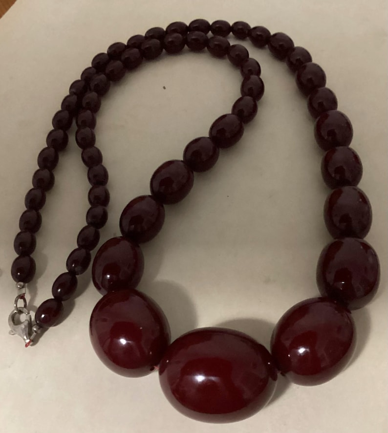 Op de afbeelding: Een ketting met gegradueerde kralen bestaande uit gepolijste, diep bordeauxrode ovale kralen. De kralen worden groter naar het midden toe, waar de grootste ovale kraal is geplaatst. De ketting wordt gesloten met een zilverkleurige metalen karabijnsluiting.