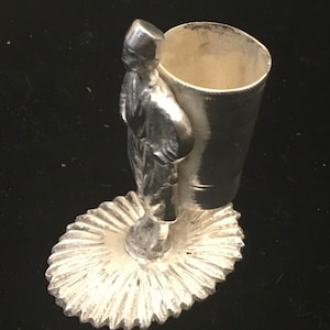 May include: A silver-colored metal figurine of a person carrying a large cylindrical container. The figure stands on a decorative, fan-shaped base. The container has a wide opening and is likely a pencil holder or similar desk accessory.