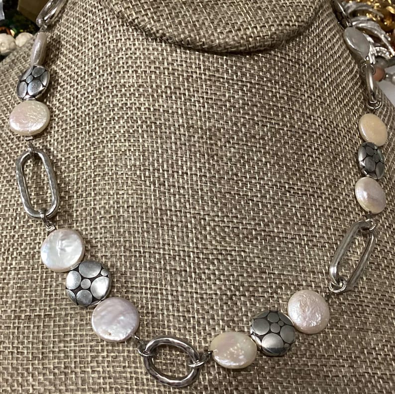May include: A necklace featuring alternating round white iridescent pearls and circular silver-toned metal discs with a textured floral pattern, connected by elongated oval silver-toned metal links in a repeating chain design.