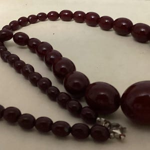 Op de afbeelding: Een ketting met gegradueerde, gepolijste, diepbordeauxrode, bolvormige kralen. De kralen worden groter naar het midden van de streng toe, die is vastgezet met een kleine, zilverkleurige metalen sluiting aan de achterkant van de ketting.