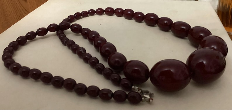 Op de afbeelding: Een ketting met gegradueerde, gepolijste, diepbordeauxrode, bolvormige kralen. De kralen worden groter naar het midden van de streng toe, die is vastgezet met een kleine, zilverkleurige metalen sluiting aan de achterkant van de ketting.
