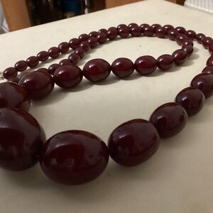 Op de afbeelding: Een ketting met gegradueerde, gepolijste, diep bordeauxrode ovale kralen. De ketting verloopt van kleinere kralen bij de sluiting naar aanzienlijk grotere, ronde kralen in het midden, verbonden door een zilverkleurige metalen sluiting.