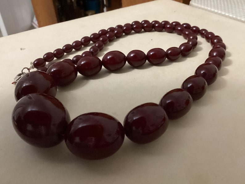 Op de afbeelding: Een ketting met gegradueerde, gepolijste, diep bordeauxrode ovale kralen. De ketting verloopt van kleinere kralen bij de sluiting naar aanzienlijk grotere, ronde kralen in het midden, verbonden door een zilverkleurige metalen sluiting.