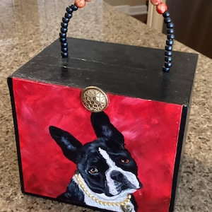 Puede incluir: Un objeto decorativo negro en forma de caja con un retrato pintado de un Boston Terrier sobre un fondo rojo. La caja tiene un asa de cuentas y un botón dorado. El perro lleva un collar dorado con un colgante de corazón.