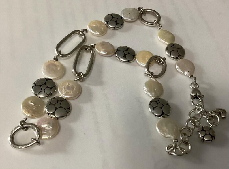 May include: A bracelet featuring alternating white coin-shaped pearls and circular silver-toned metal discs with a dotted pattern, connected by silver-toned oval links. The piece includes a silver-toned toggle clasp and an adjustable chain extension.