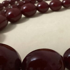Op de afbeelding: Een ketting met een streng gladde, gepolijste, diepbordeauxrode kralen. De kralen zijn in grootte gegradueerd, met grotere, afgeronde, ovale kralen aan de voorzijde en kleinere, ronde kralen langs de rest van de streng.