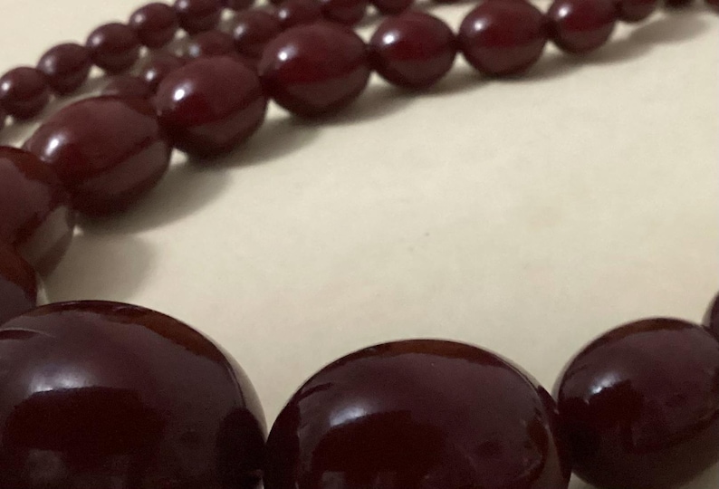 Op de afbeelding: Een ketting met een streng gladde, gepolijste, diepbordeauxrode kralen. De kralen zijn in grootte gegradueerd, met grotere, afgeronde, ovale kralen aan de voorzijde en kleinere, ronde kralen langs de rest van de streng.