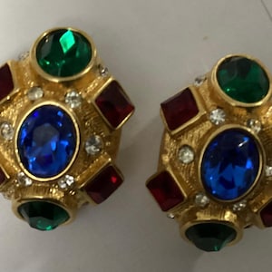 May include: A pair of gold-tone earrings featuring a central oval blue gemstone, surrounded by four square red gemstones, two round green gemstones, and small clear accent crystals set into a textured gold-colored metal base.