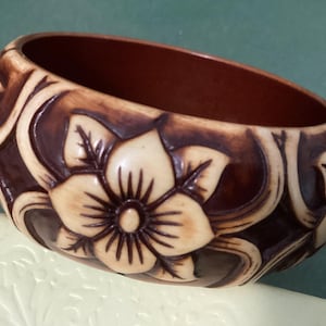 May include: A carved wooden bangle bracelet with a floral design. The bracelet is primarily brown with cream-colored floral accents. The flowers and leaves are carved into the surface, creating a textured appearance. The interior of the bracelet is a darker brown.