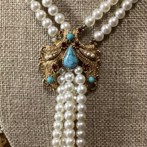 May include: A multi-strand necklace featuring white faux pearls and a central gold-tone metal pendant. The pendant is decorated with a teardrop-shaped turquoise stone, small round turquoise accents, and deep red inset stones in an ornate setting.