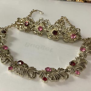 May include: A gold-tone metal necklace featuring a series of repeating floral-inspired links. Each link is textured with fine lines and accented with round, faceted rhinestones in varying shades of pink and deep red, secured by a metal hook clasp.