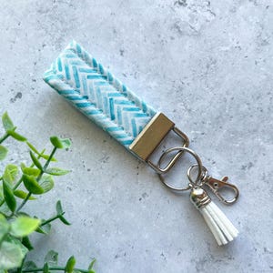 May include: A wristlet keychain with a light blue and white chevron pattern. The keychain has a gold-colored metal clasp and a silver keyring with a white tassel. The wristlet is approximately 6 inches long.