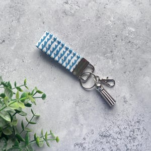 May include: A blue and white patterned wristlet keychain with a silver-colored metal clasp and tassel. The wristlet has a repeating wave-like design. The keychain is on a gray surface with green foliage in the corner.