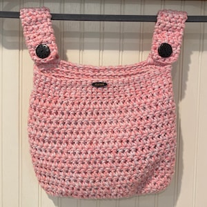 May include: A pink and white crocheted baby bib with two straps and black buttons. The bib has a small tag that says "Hand Made".