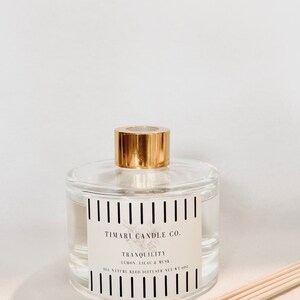 May include: A clear glass diffuser bottle with a gold lid and a black and white striped label. The label reads "TIMARI CANDLE CO. TRANQUILITY LEMON, LILAC & MUSK ALL NATURAL REED DIFFUSER NET WT 4 OZ". The bottle is sitting on a beige surface with a set of wooden diffuser sticks.