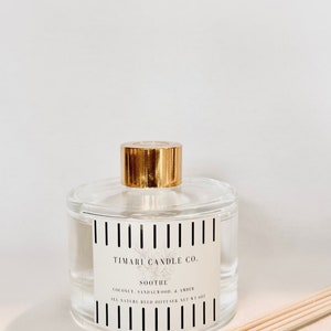 May include: A clear glass bottle with a gold lid and a black and white striped label. The label reads "TIMARI CANDLE CO. SOOTHE COCONUT, SANDALWOOD, & AMBER ALL NATURAL REED DIFFUSER NET WT 6 OZ". The bottle is sitting on a beige surface with wooden sticks in front of it.
