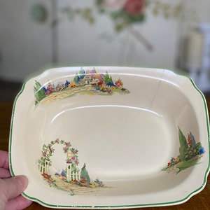 May include: A white oval serving dish with a green rim and a floral design. The design features a cottage with a path leading to a white picket fence with a rose archway.