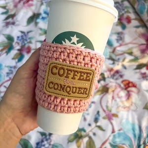 Op de afbeelding: Een roze gehaakte koffiebekerwarmer met een bruin leren patch waarop "Coffee & Conquer" staat.