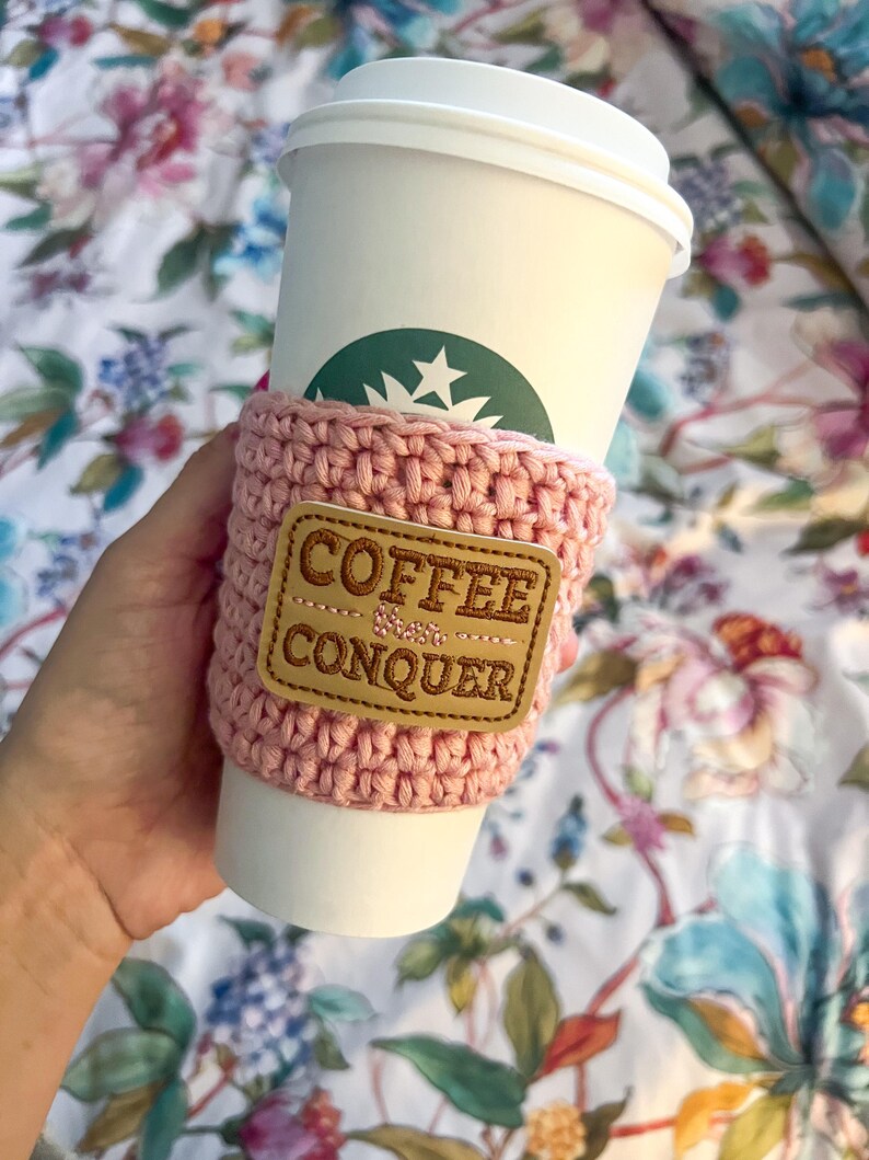 Op de afbeelding: Een roze gehaakte koffiebekerwarmer met een bruin leren patch waarop "Coffee & Conquer" staat.