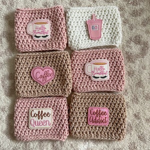 Op de afbeelding: Zes gehaakte koffiebekers in roze, bruin en wit met verschillende geborduurde ontwerpen, waaronder "Hello Beautiful", "Coffee", "Coffee Queen" en "Coffee Addict".
