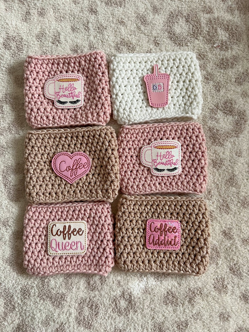 Op de afbeelding: Zes gehaakte koffiebekers in roze, bruin en wit met verschillende geborduurde ontwerpen, waaronder "Hello Beautiful", "Coffee", "Coffee Queen" en "Coffee Addict".