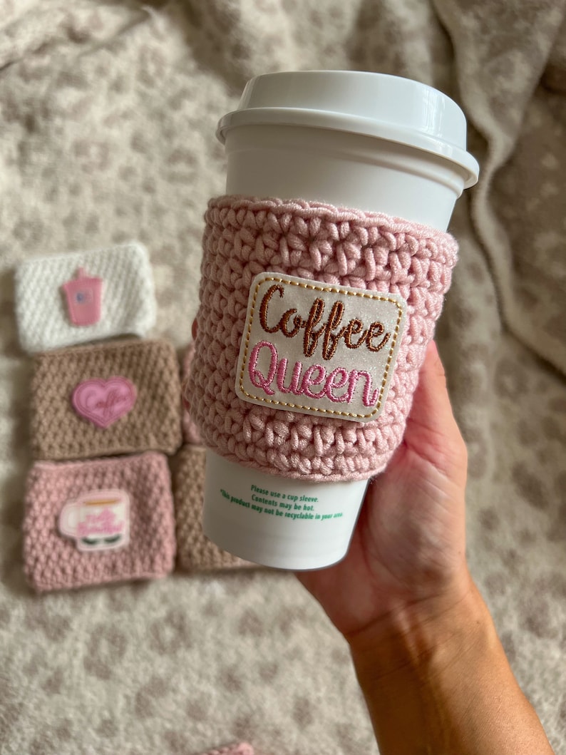 Op de afbeelding: Roze gebreide koffiebekerhoes met een gouden geborduurde patch met de tekst "Coffee Queen".