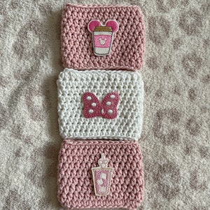 Op de afbeelding: Drie gehaakte koffiebekers in roze en wit met verschillende geborduurde ontwerpen. De ontwerpen omvatten een roze koffiebeker met Mickey Mouse-oren, een roze strik met witte stippen en een roze koffiebeker met een rietje.