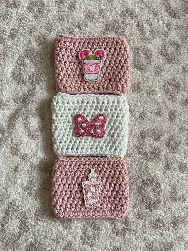 Op de afbeelding: Drie gehaakte koffiebekers in roze en wit met verschillende geborduurde ontwerpen. De ontwerpen omvatten een roze koffiebeker met Mickey Mouse-oren, een roze strik met witte stippen en een roze koffiebeker met een rietje.
