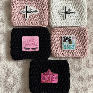 Op de afbeelding: Vijf gehaakte koffiebekers met verschillende geborduurde patches. De bekers zijn roze, wit en zwart. De patches bevatten de woorden "faith", "hello beautiful", "bee kind" en "mother hustler".