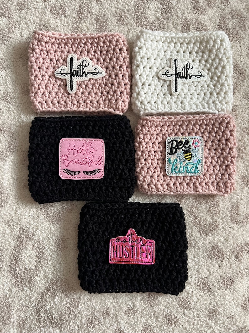 Op de afbeelding: Vijf gehaakte koffiebekers met verschillende geborduurde patches. De bekers zijn roze, wit en zwart. De patches bevatten de woorden "faith", "hello beautiful", "bee kind" en "mother hustler".