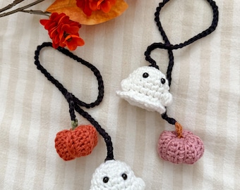 Puede incluir: Dos adornos de Halloween hechos a mano con ganchillo. Uno presenta un fantasma blanco con ojos negros y una calabaza naranja. El otro tiene un fantasma blanco y una calabaza rosa. Ambos tienen lazos negros para colgar.
