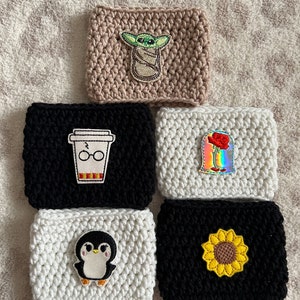 Op de afbeelding: Zes gehaakte koffiebekers met verschillende geborduurde ontwerpen. De bekers zijn beige, zwart en wit. De ontwerpen omvatten een baby Yoda, een koffiebeker met een bril, een roos, een pingu&iuml;n en een zonnebloem.