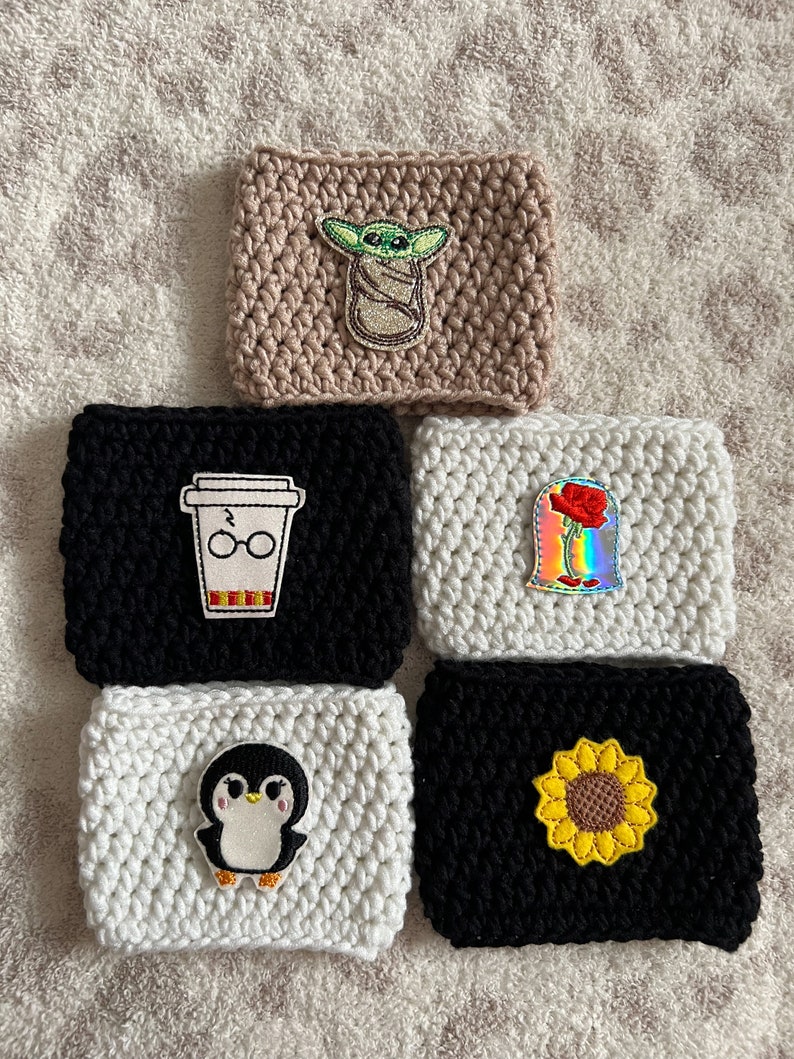 Op de afbeelding: Zes gehaakte koffiebekers met verschillende geborduurde ontwerpen. De bekers zijn beige, zwart en wit. De ontwerpen omvatten een baby Yoda, een koffiebeker met een bril, een roos, een pingu&iuml;n en een zonnebloem.