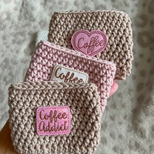 Op de afbeelding: Drie gehaakte koffiebekers in roze en beige tinten. De bekers hebben roze viltpatches met de woorden "Coffee", "Coffee" en "Coffee Addict" in gouden letters.