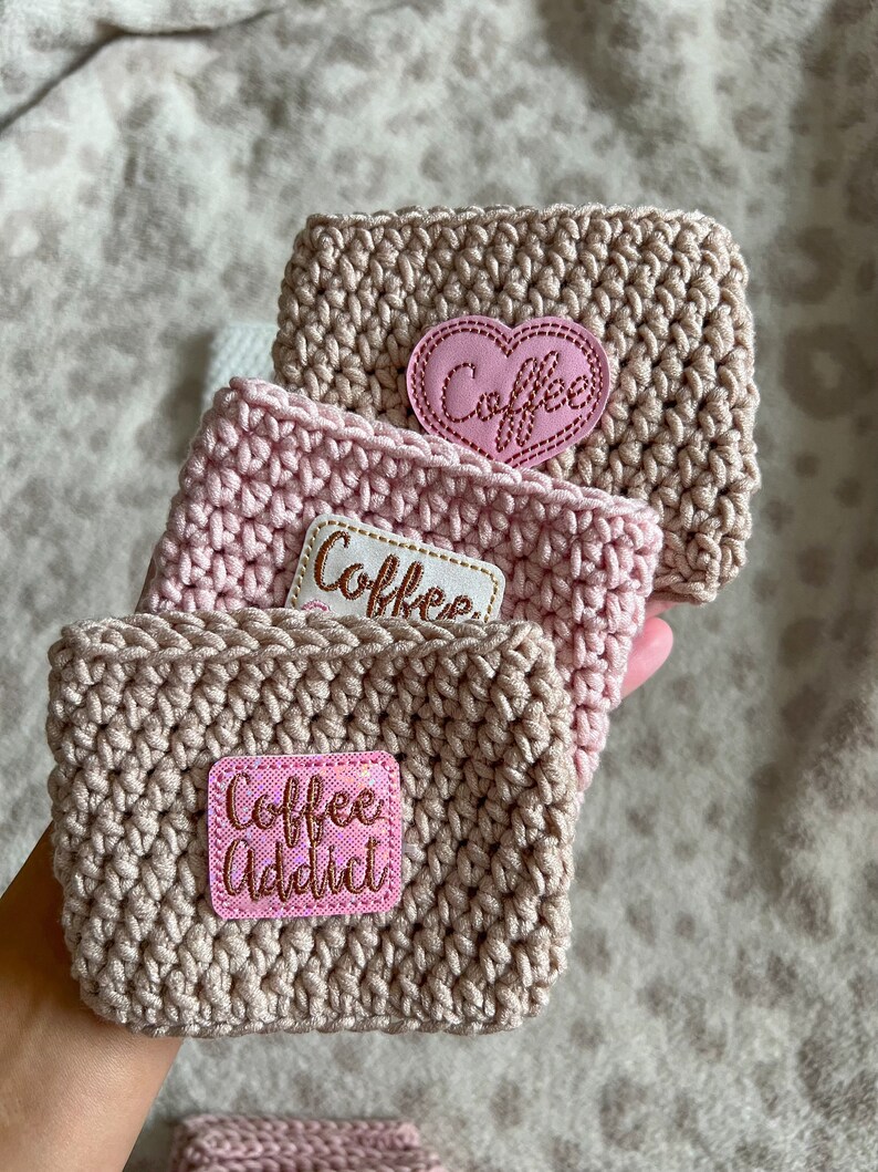 Op de afbeelding: Drie gehaakte koffiebekers in roze en beige tinten. De bekers hebben roze viltpatches met de woorden "Coffee", "Coffee" en "Coffee Addict" in gouden letters.