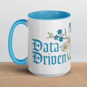 Peut inclure: Mug en céramique blanche avec intérieur et anse bleus. Le mug présente le texte "Data Driven" en bleu, accompagné d'illustrations florales bleues et blanches. Le mug est posé sur une surface en bois.
