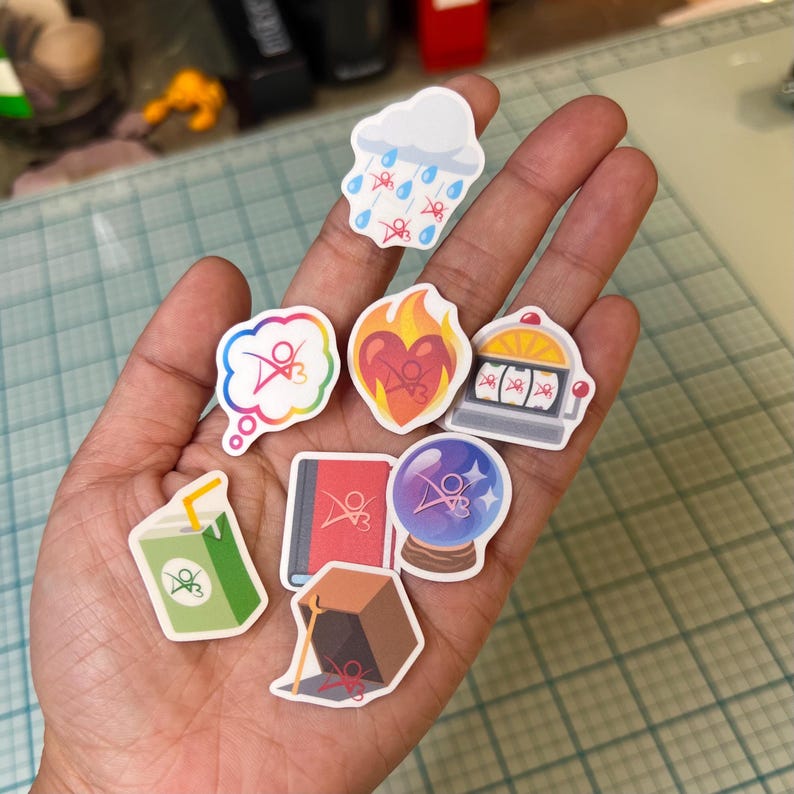May include: A hand holding nine colorful stickers with various designs, including a cloud with raindrops, a heart with flames, a slot machine, a book, a crystal ball, a thought bubble, a box, and a juice box. Each sticker features a stylized logo with a red, white, and blue design.