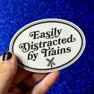 May include: An oval sticker with the text "Easily Distracted by Trains" in black script. The sticker has a black border and a railroad crossing symbol. The background is a sparkly blue.