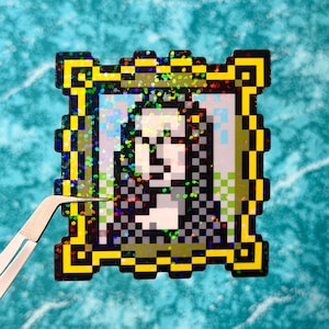 May include: A pixelated sticker featuring the Mona Lisa in a gold frame. The artwork is rendered in a retro, 8-bit style with a holographic, glittery finish. A pair of tweezers is holding the sticker.