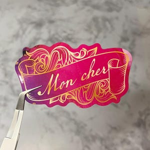 May include: A pink and purple sticker with gold foil accents. The sticker features a scroll with the text "Mon cher" in gold lettering.