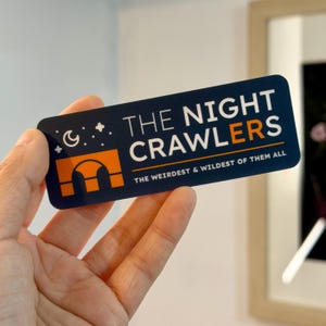 May include: A rectangular sticker with rounded corners, held in a hand. The sticker features the text "THE NIGHT CRAWLERS" in white, with an orange graphic and the tagline "THE WEIRDEST & WILDEST OF THEM ALL" in white. The background is navy blue.
